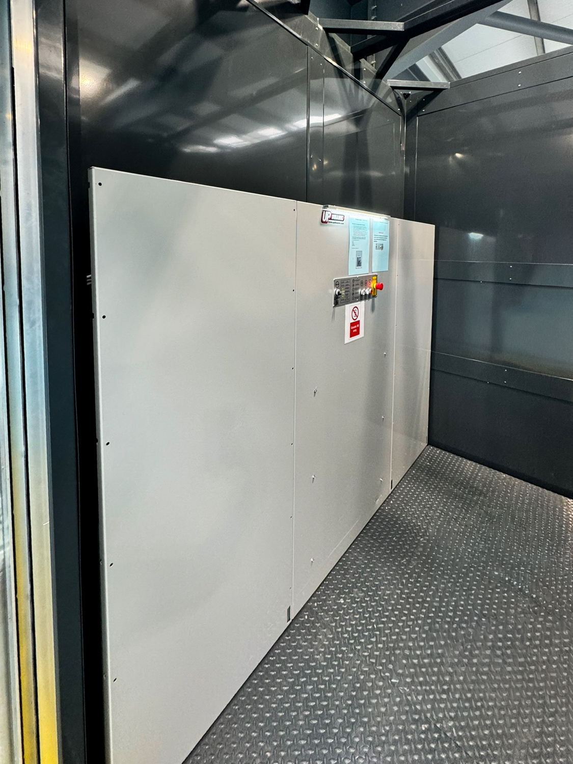 Interior of a metal goods lift with gray panel walls and a control panel with buttons and warning signs.