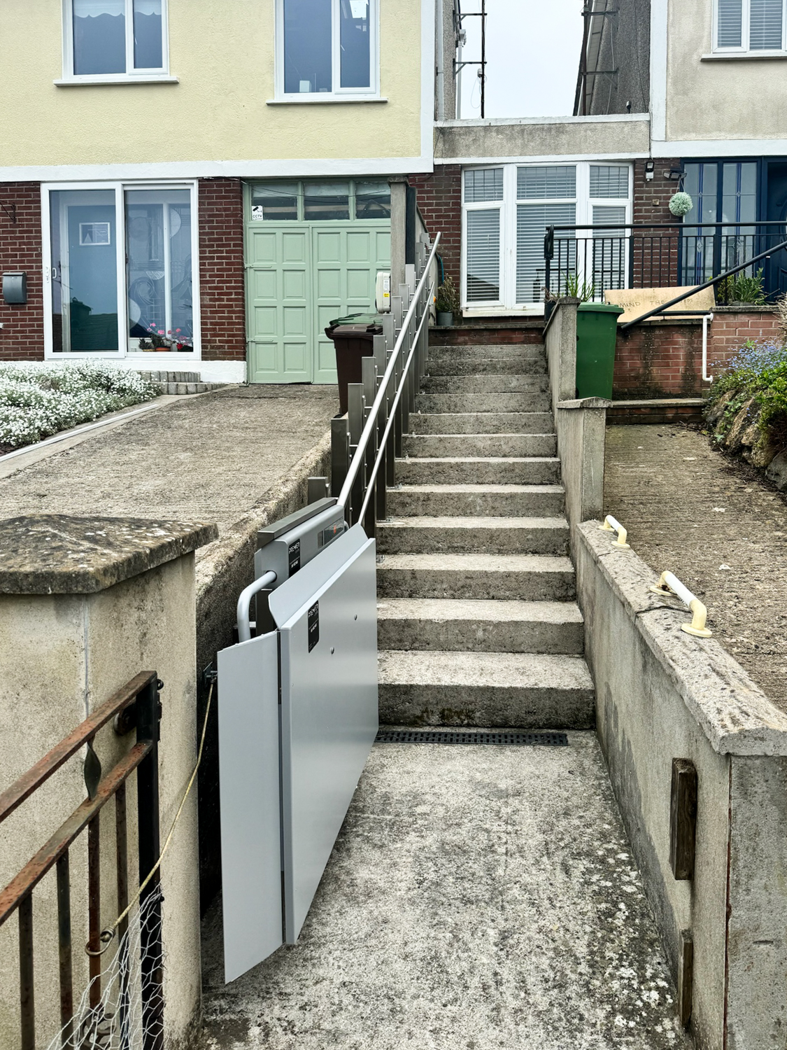 Outdoor concrete staircase with a metal accessibility wheelchair lift installed alongside it leading up to residential houses.