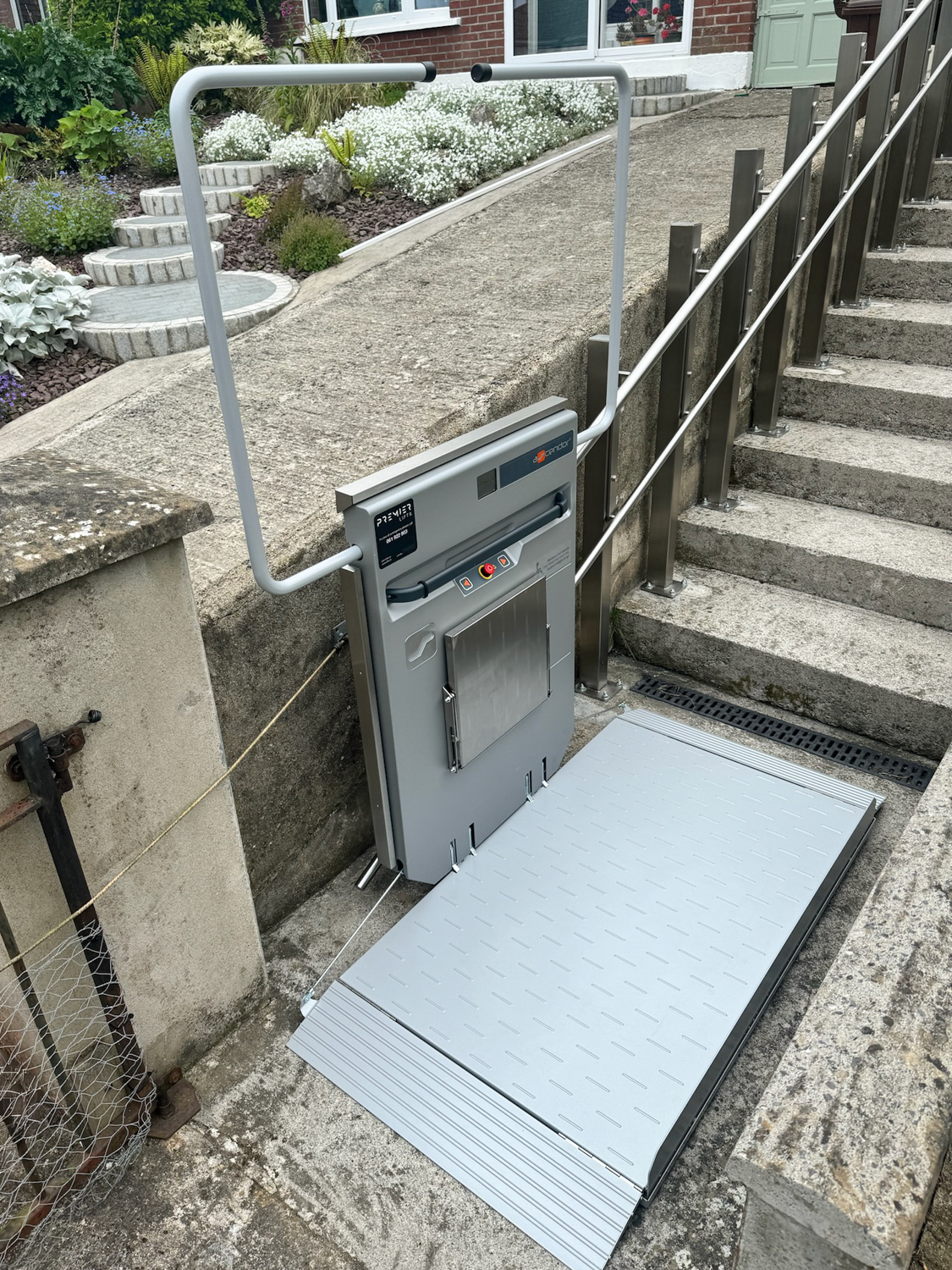 Outdoor wheelchair lift platform installed next to concrete stairs with handrails and a garden in the background.