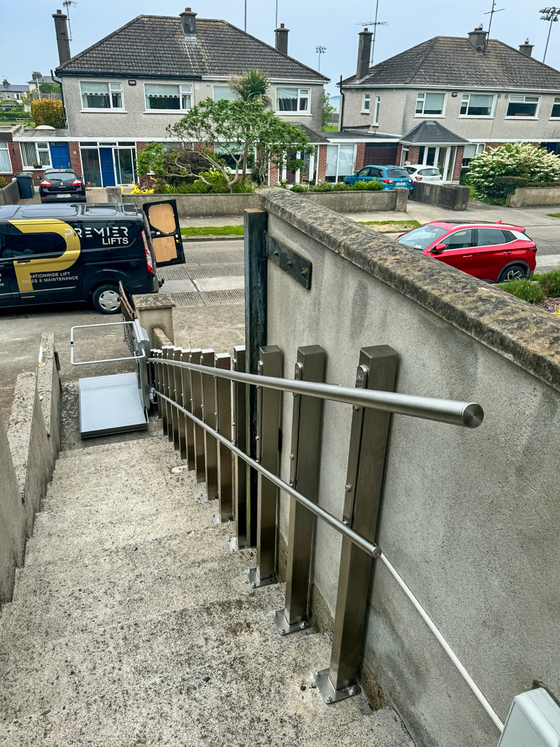 Concrete outdoor stairs with a metal handrail and a wheelchair lift at the bottom, residential houses and parked cars in the background.