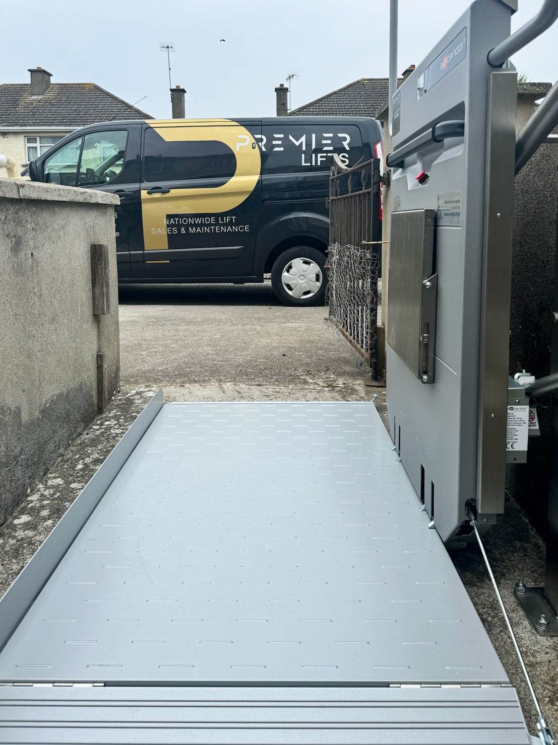 Metal wheelchair platform lift installed on a concrete ramp outdoors, with a Premier Lifts van parked in the background.