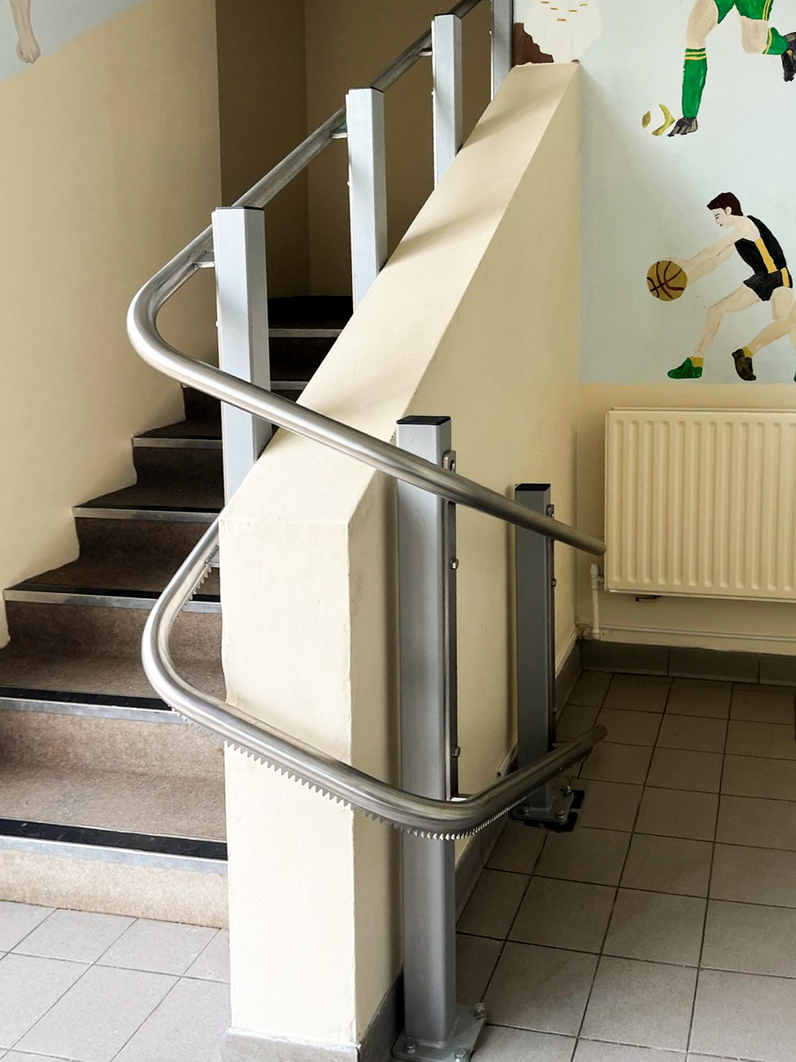 Curved metal stairlift rail installed along a stairway with sports-themed wall mural in background.