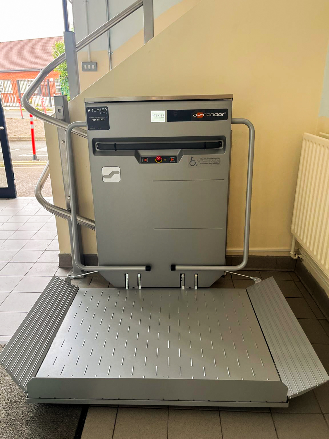 Indoor silver Ascendor wheelchair platform lift installed beside a staircase with control buttons and safety rails.
