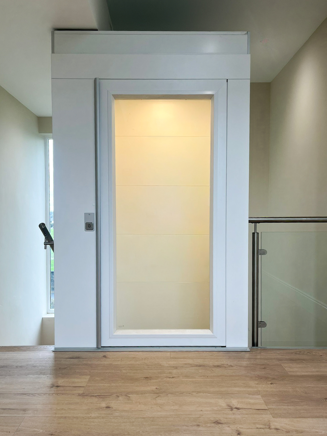 White public platform lift with glass door in a modern hallway with wooden flooring.