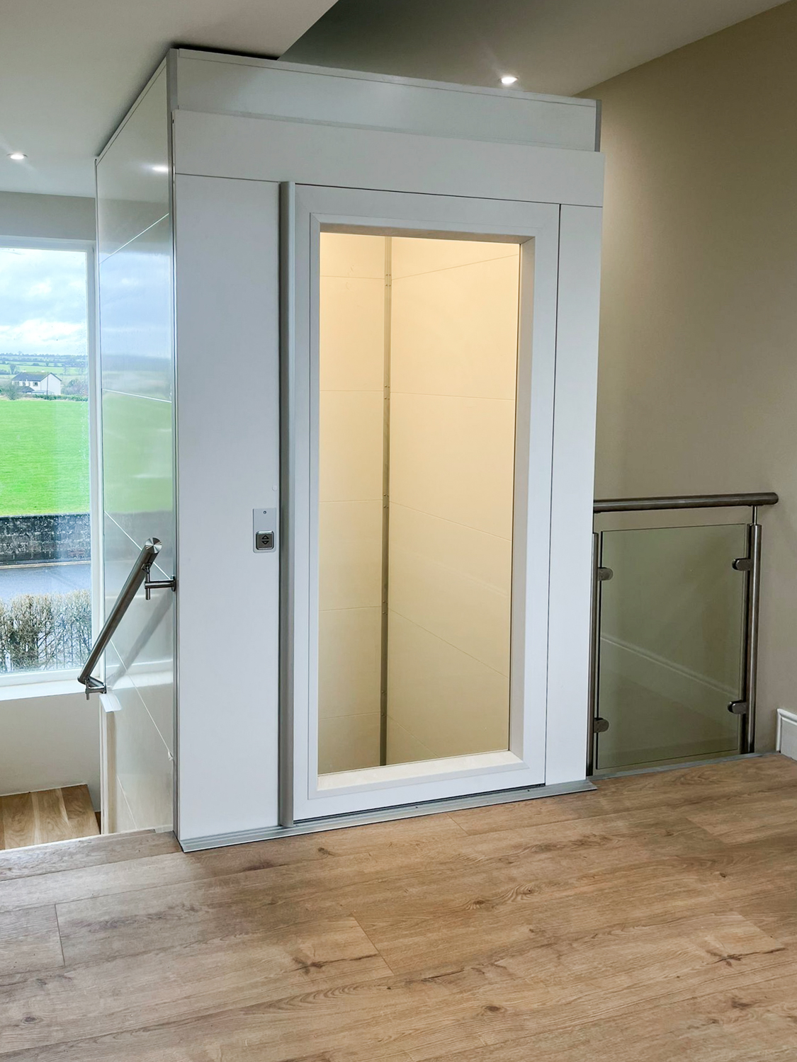 Indoor platform lift with a glass door, located at the top of a stairway with wooden flooring.