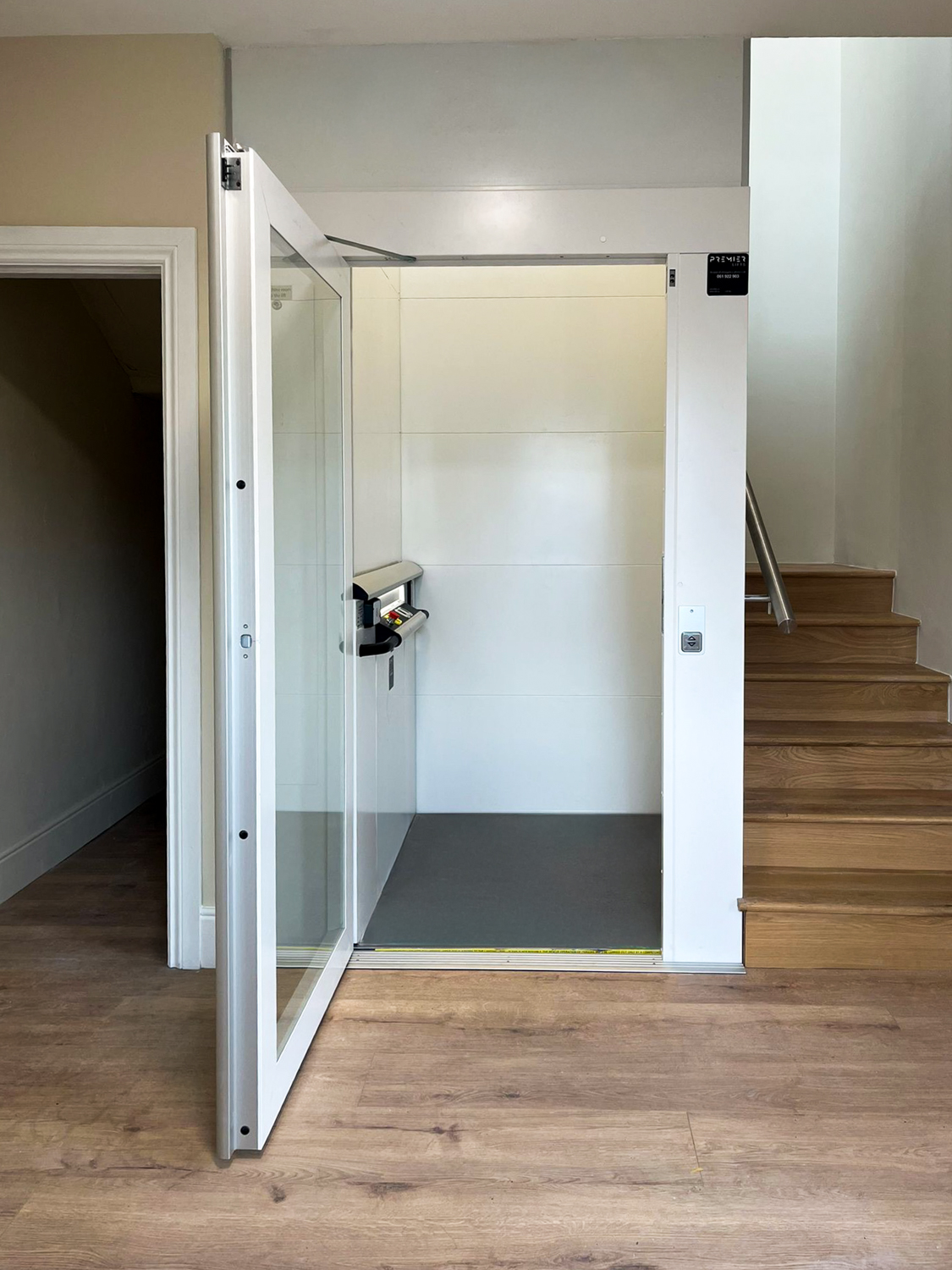 Open white platform lift beside wooden stairs inside a building with light-colored walls and wood flooring.