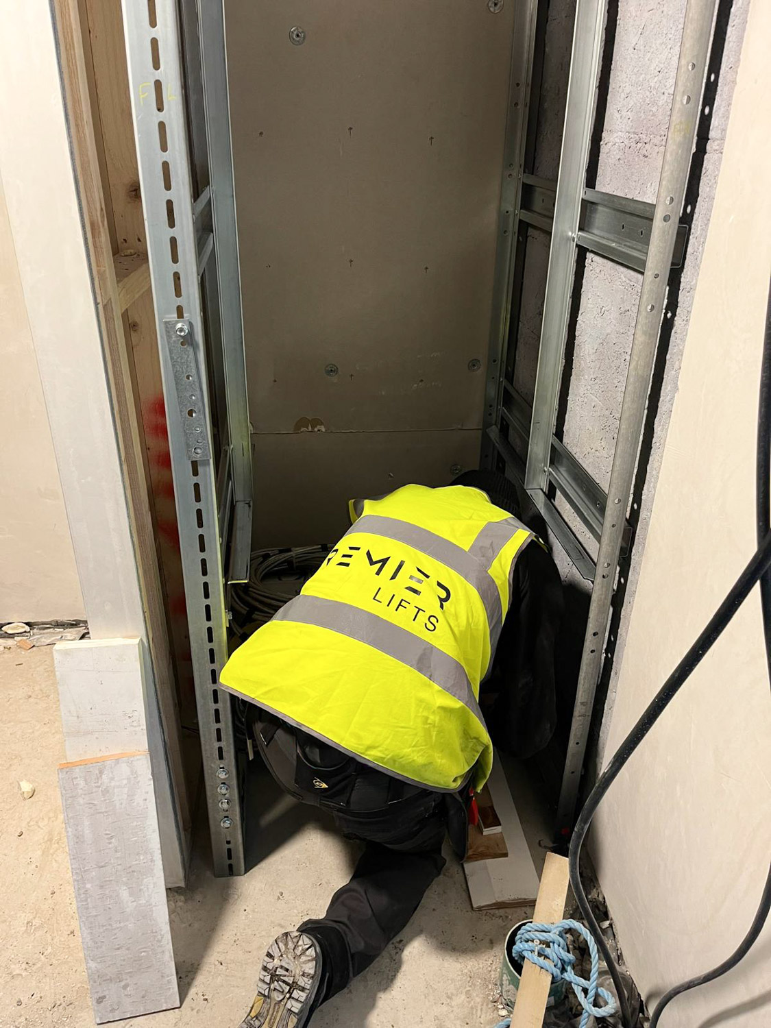 Worker in a yellow Premier Lifts safety vest kneeling and inspecting installation inside a small lift shaft.