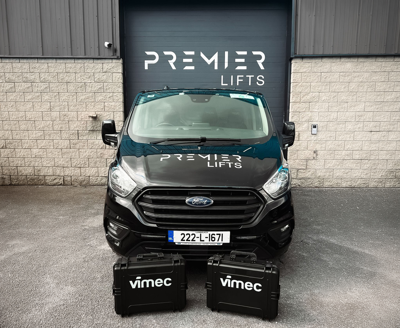 Front view of a black Ford van with Premier Lifts branding parked in front of a black garage door with the same logo, two black Vimec cases on the ground.