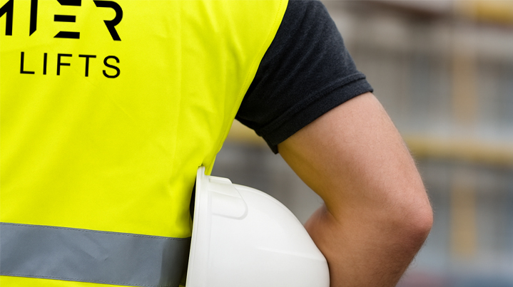 Worker wearing a yellow high-visibility vest with 'LIFTS' printed on it, holding a white safety helmet under their arm.