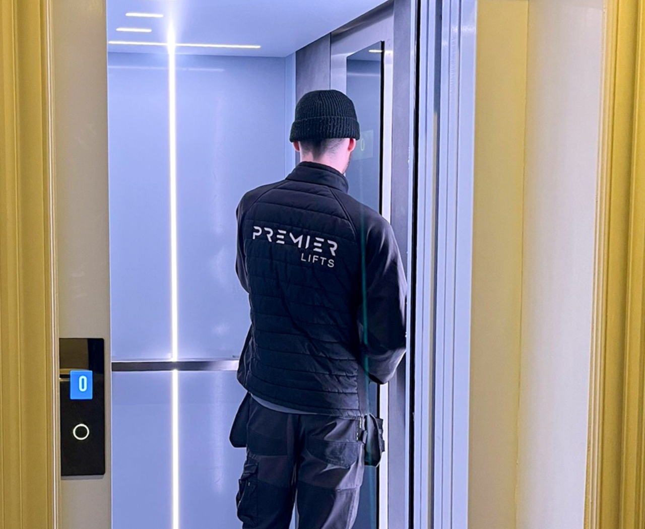Technician wearing a Premier Lifts jacket standing in an open elevator with illuminated floor indicator showing 0.