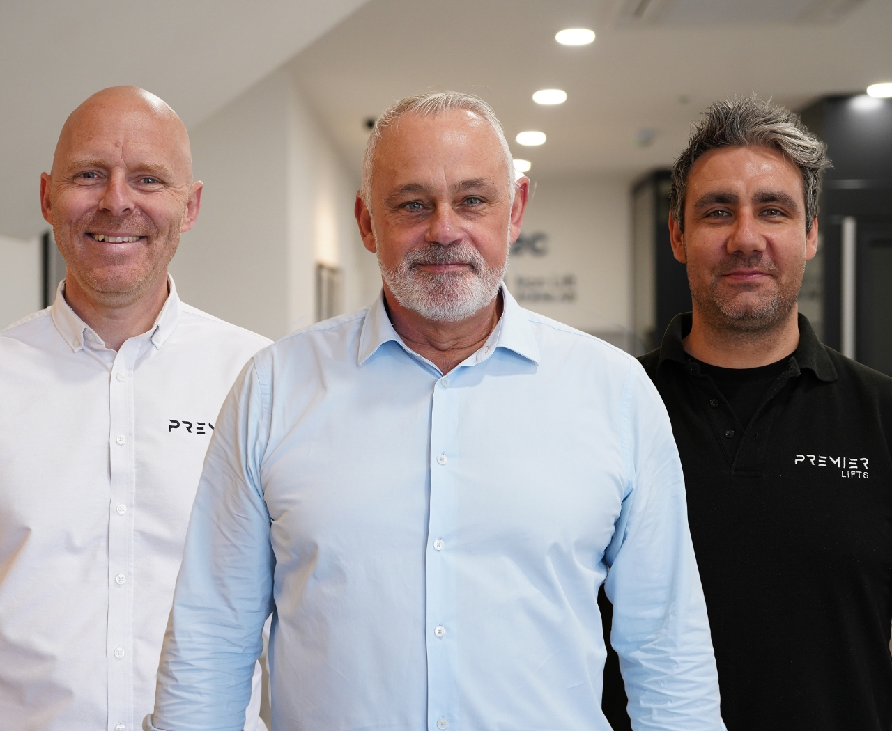 Three men standing indoors, two wearing Premier Lifts branded shirts in white and black, and one in a light blue shirt.