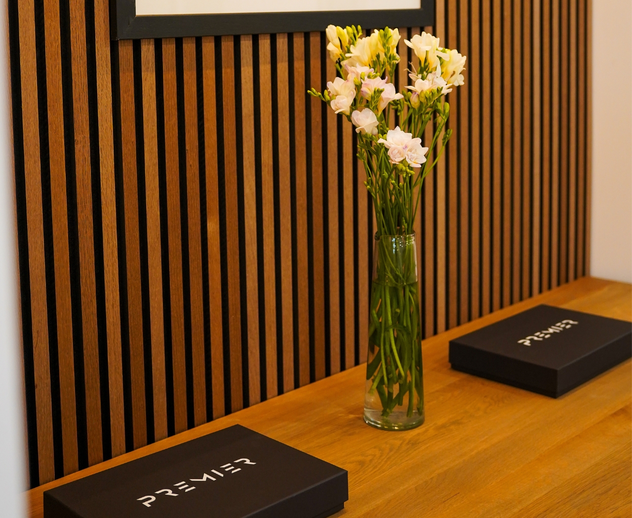 Glass vase with white and pale pink flowers on a wooden table with two black boxes labeled 'Premier Lifts' in front of a vertical wood slat wall.