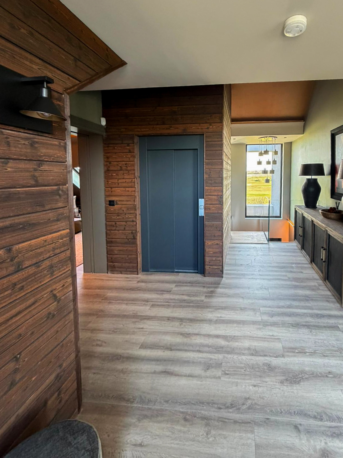 Modern interior hallway with wooden walls, a closed blue elevator door, light wood flooring, and a large window overlooking a green landscape.