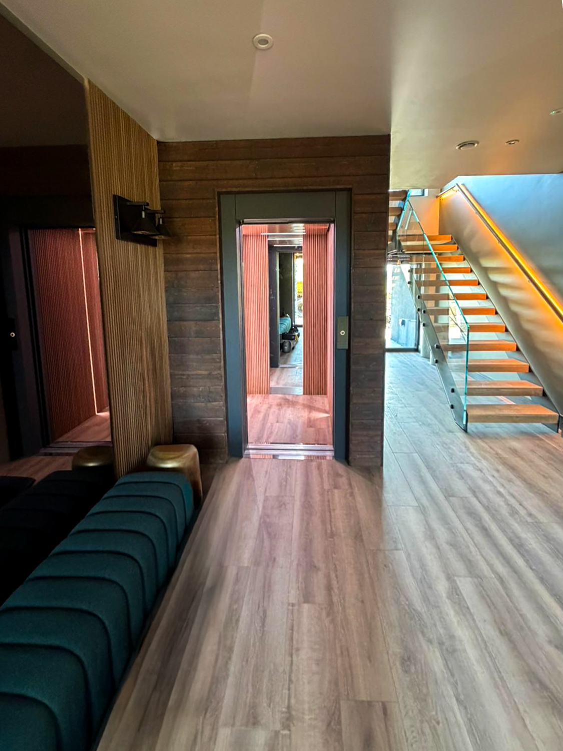 Modern interior with a wooden floor, glass-paneled floating stairs with yellow lighting along the handrail, green cushioned bench, and an open elevator with wood paneling inside.