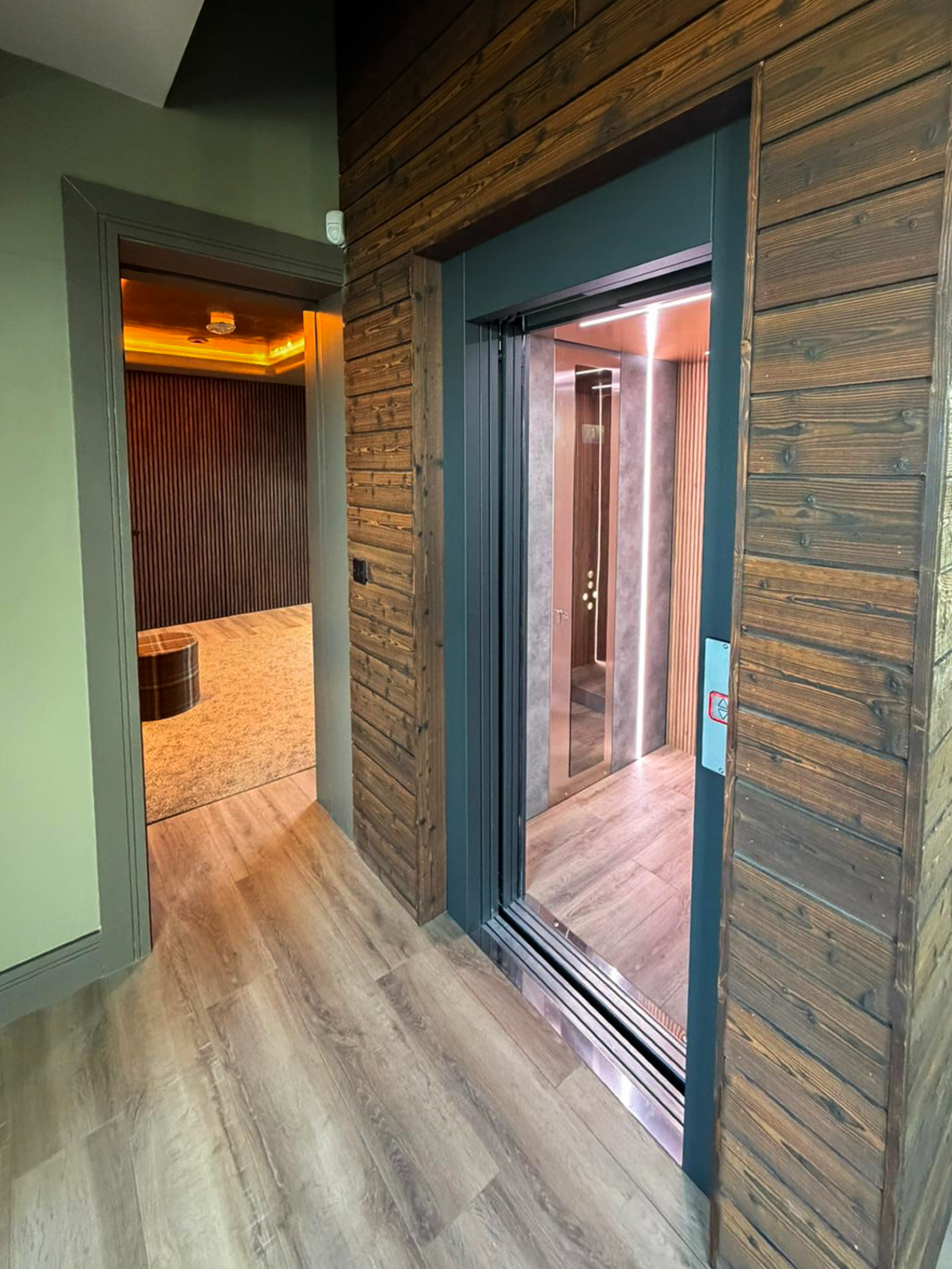 Open modern elevator with wooden paneling inside, next to a hallway leading to a room with warm lighting and wooden slat wall.
