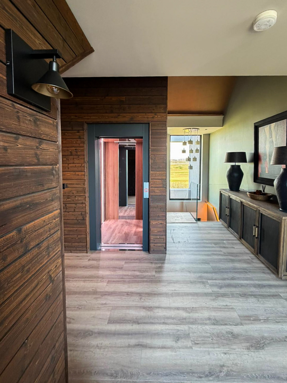 Modern hallway with light wood flooring, wooden wall paneling, an open elevator with reflective interior, and a sideboard with two black lamps and a framed picture.