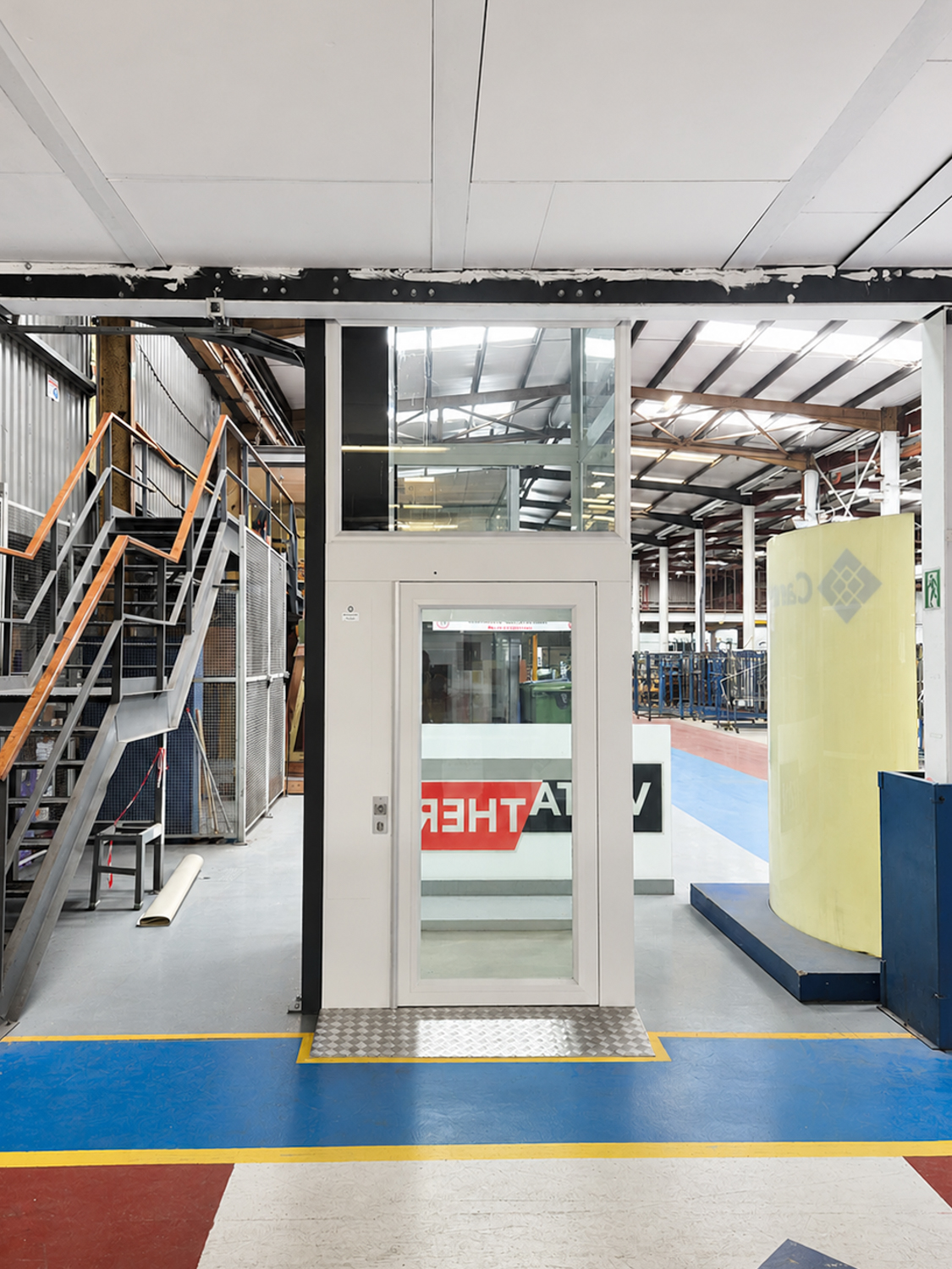 Modern white indoor platform lift with glass panels in a factory space near metal stairs.