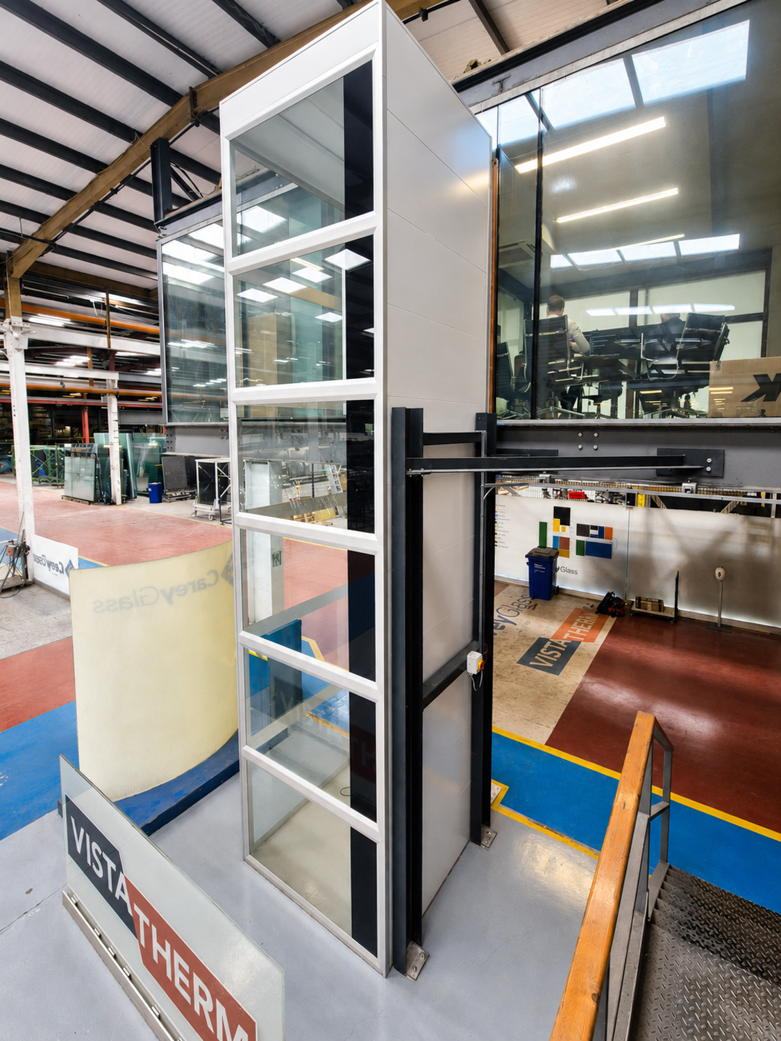 Glass platform lift in industrial warehouse with office meeting room above.