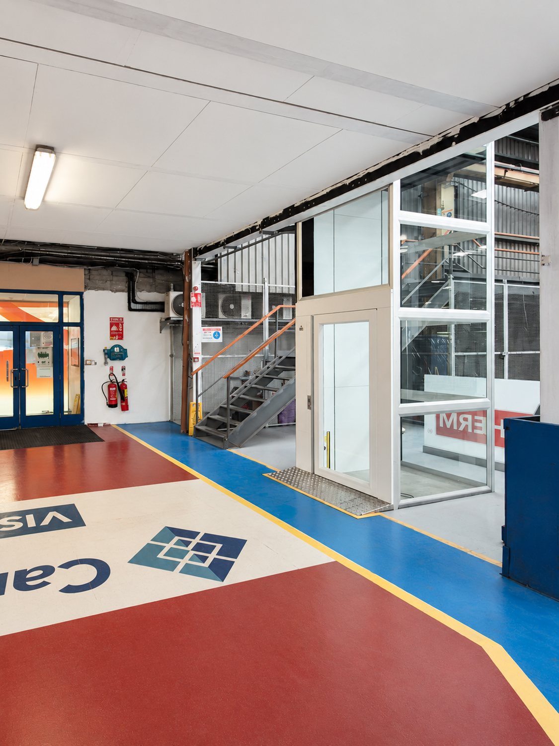 Indoor industrial space with glass platform lift, blue and red flooring, and metal staircase.