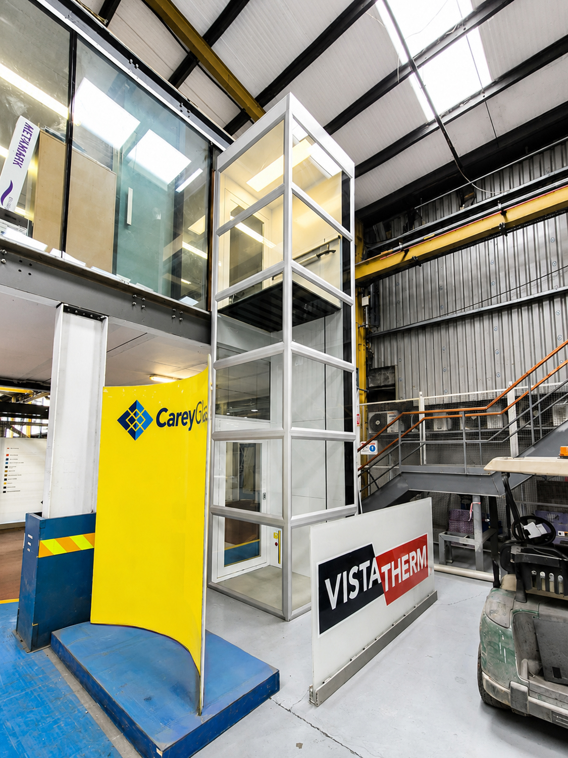 Indoor glass public platform lift with metal frame between yellow CareyGlass and white Vistatherm signs.