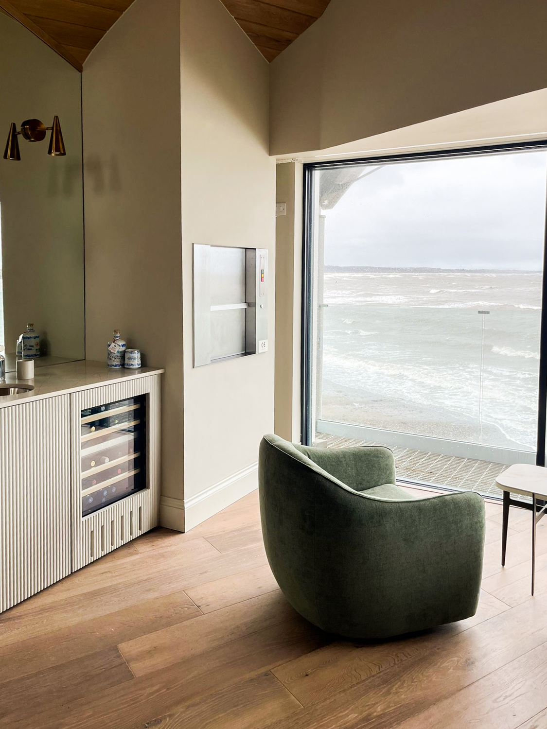 Green armchair facing large window overlooking a rough sea, next to a cabinet with wine fridge and a dumbwaiter lift.