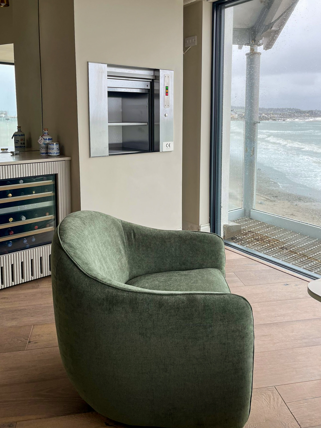 Green armchair in modern room with glass dumbwaiter lift, wine fridge, and seaside view through window.