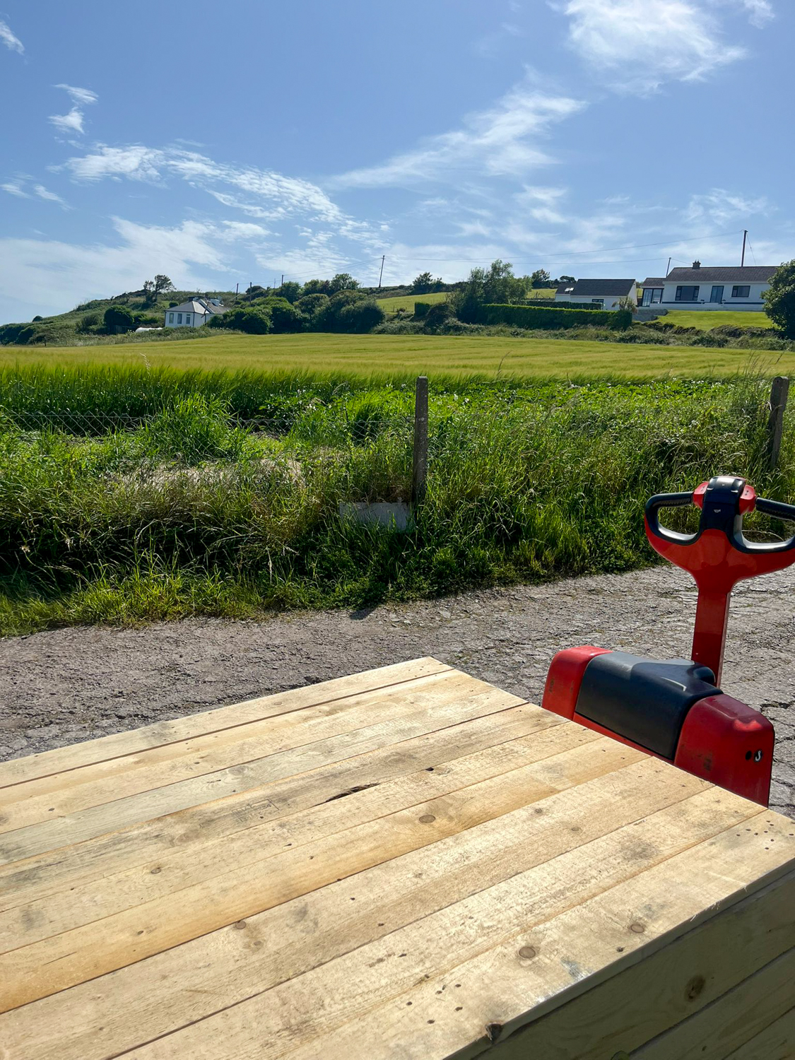 Red pallet jack on a dirt road next to a wooden pallet with green fields and houses in the background.