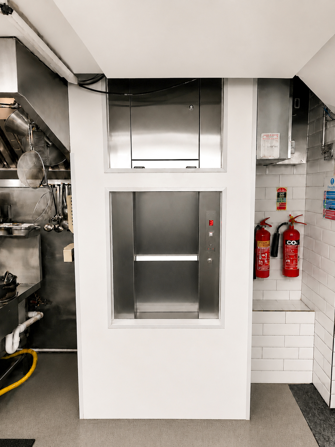 Stainless steel dumbwaiter lift in a kitchen with fire extinguishers on tiled wall.