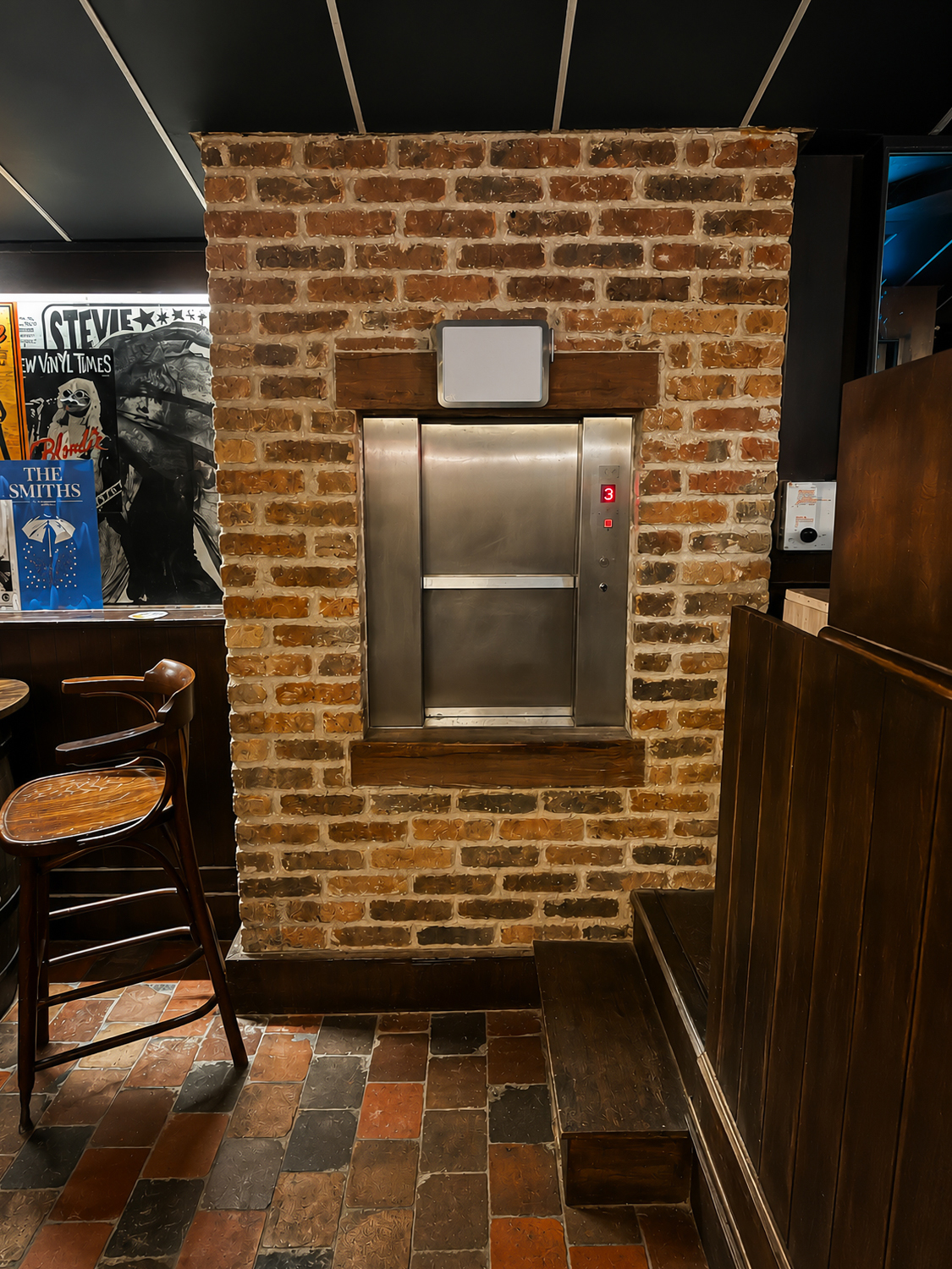 Stainless steel dumbwaiter lift set in a brick wall in a pub with a wooden chair and steps nearby.