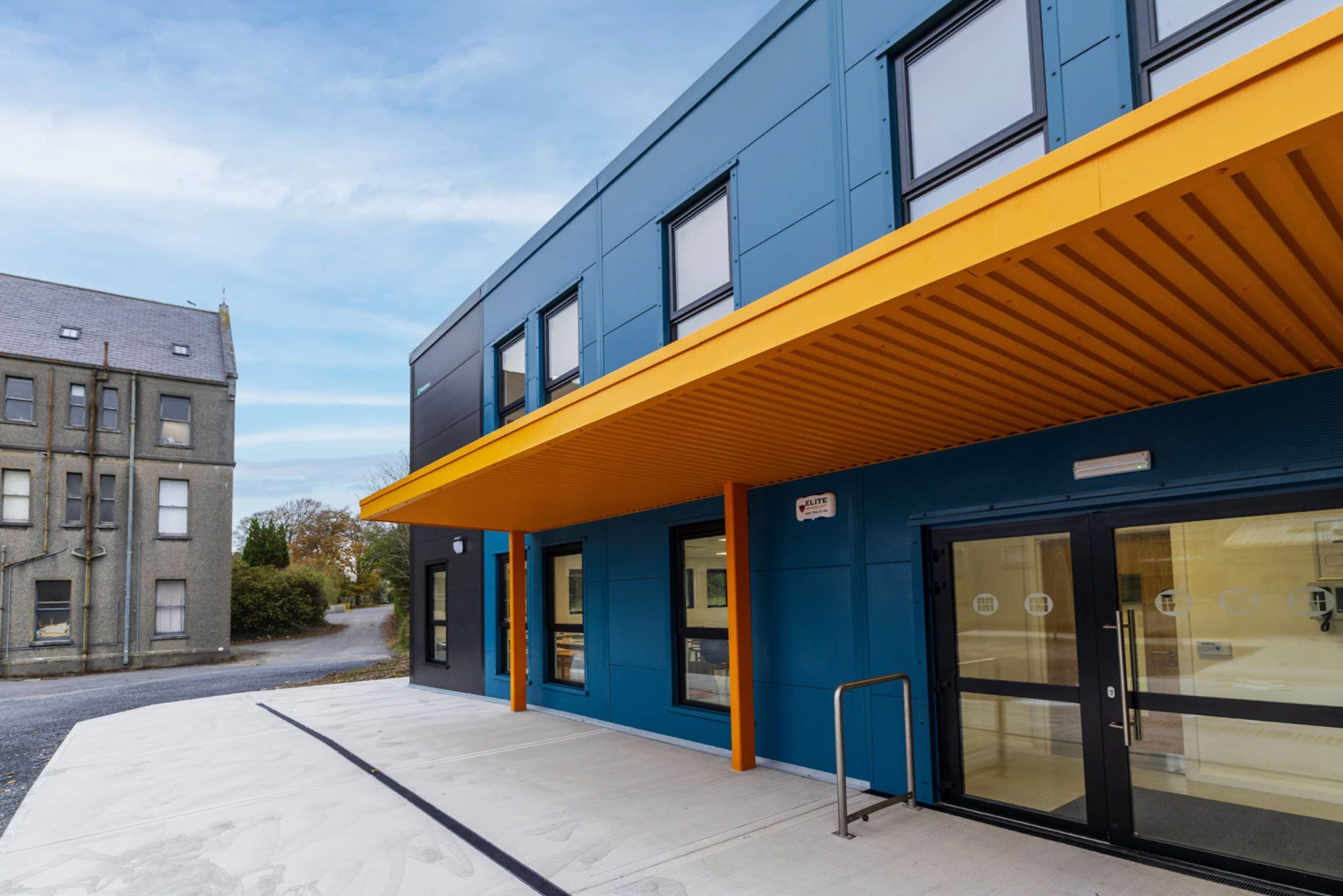 Modern blue building with yellow canopy and glass entrance doors beside an older gray building.