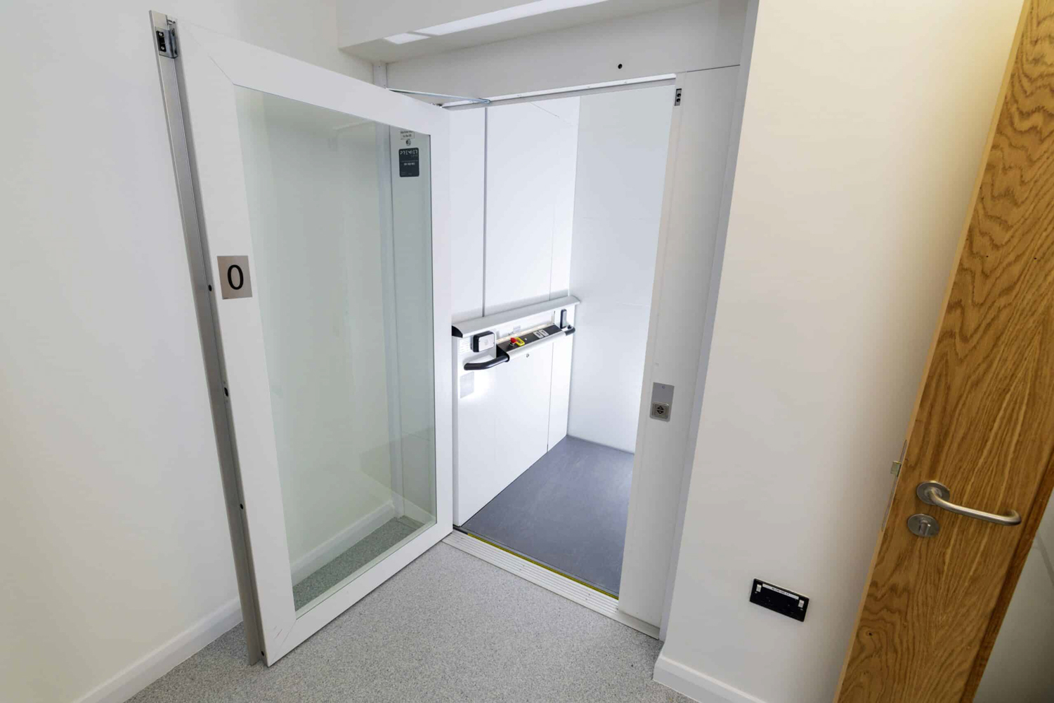 Open white vertical public platform lift door in hallway with wooden door to the side.