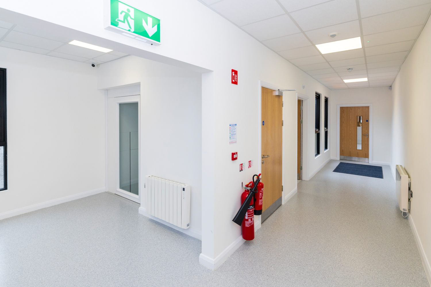 Bright hallway with wooden doors, fire extinguishers, exit sign, gray flooring, and white walls.