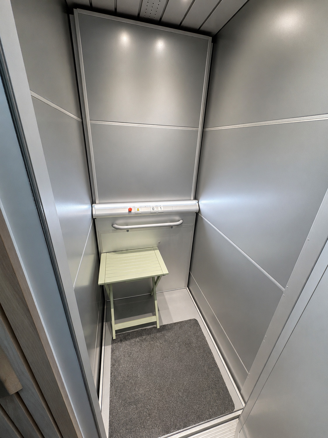 Interior of home platform lift with gray metallic walls, safety rail, control buttons, and a folding chair inside.