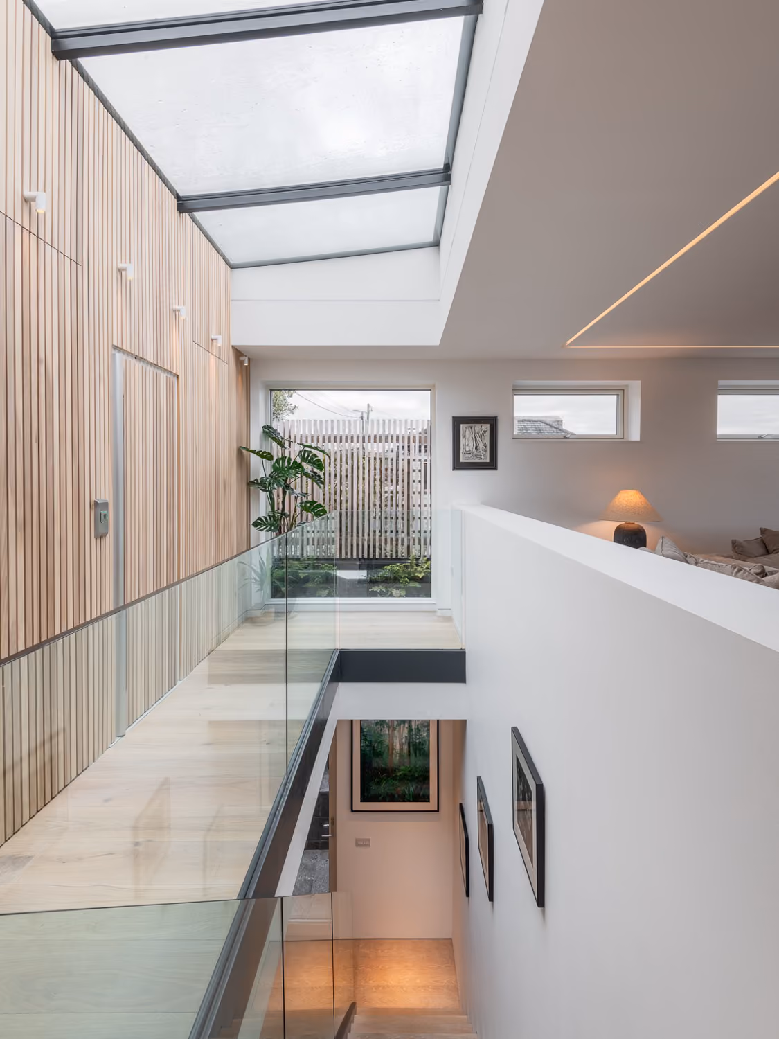 Modern indoor stairway with glass railings, wooden wall paneling, skylight, and plants near the window.