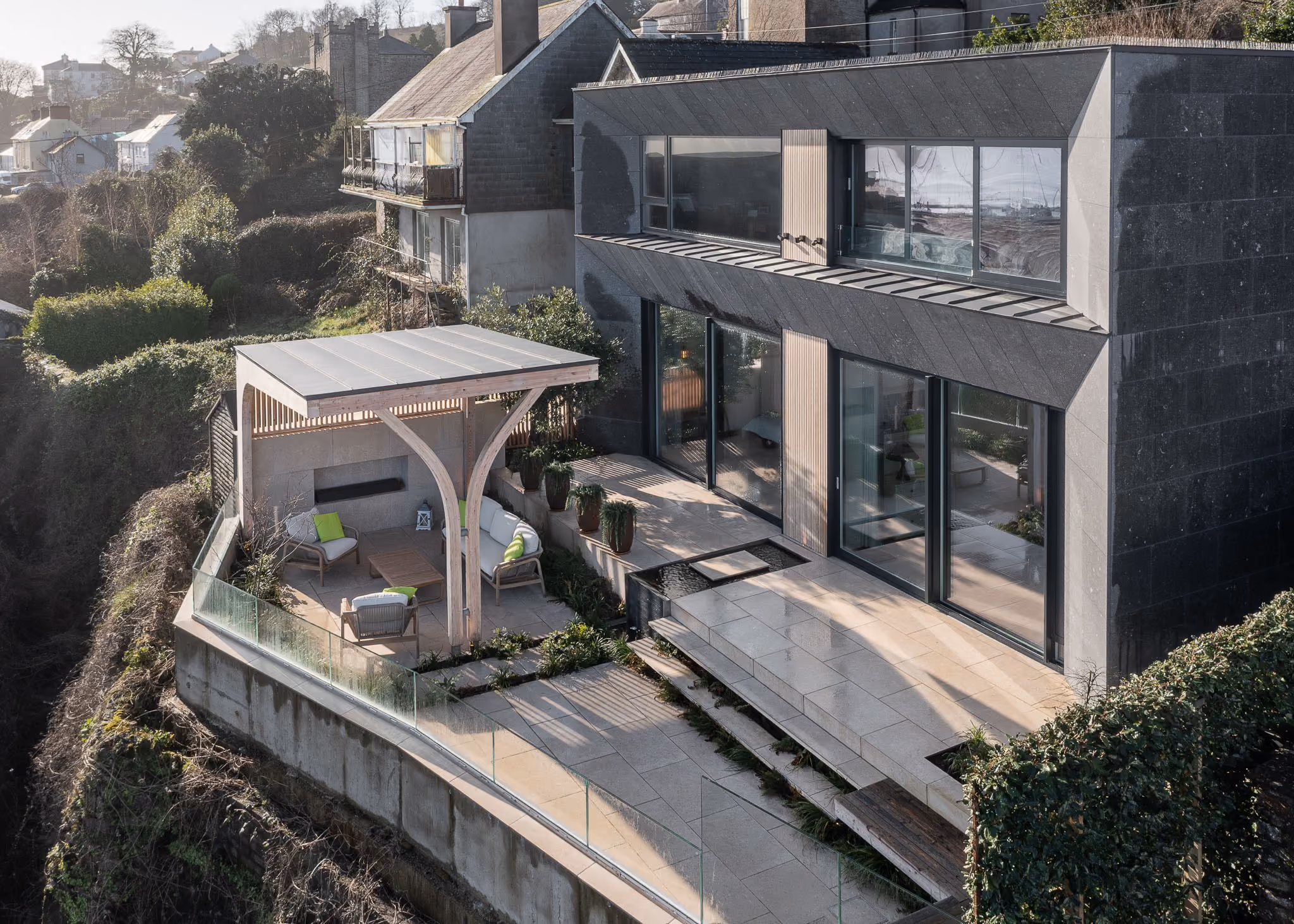 Modern two-story house with large windows, outdoor seating area, glass railing, and wooden pergola.