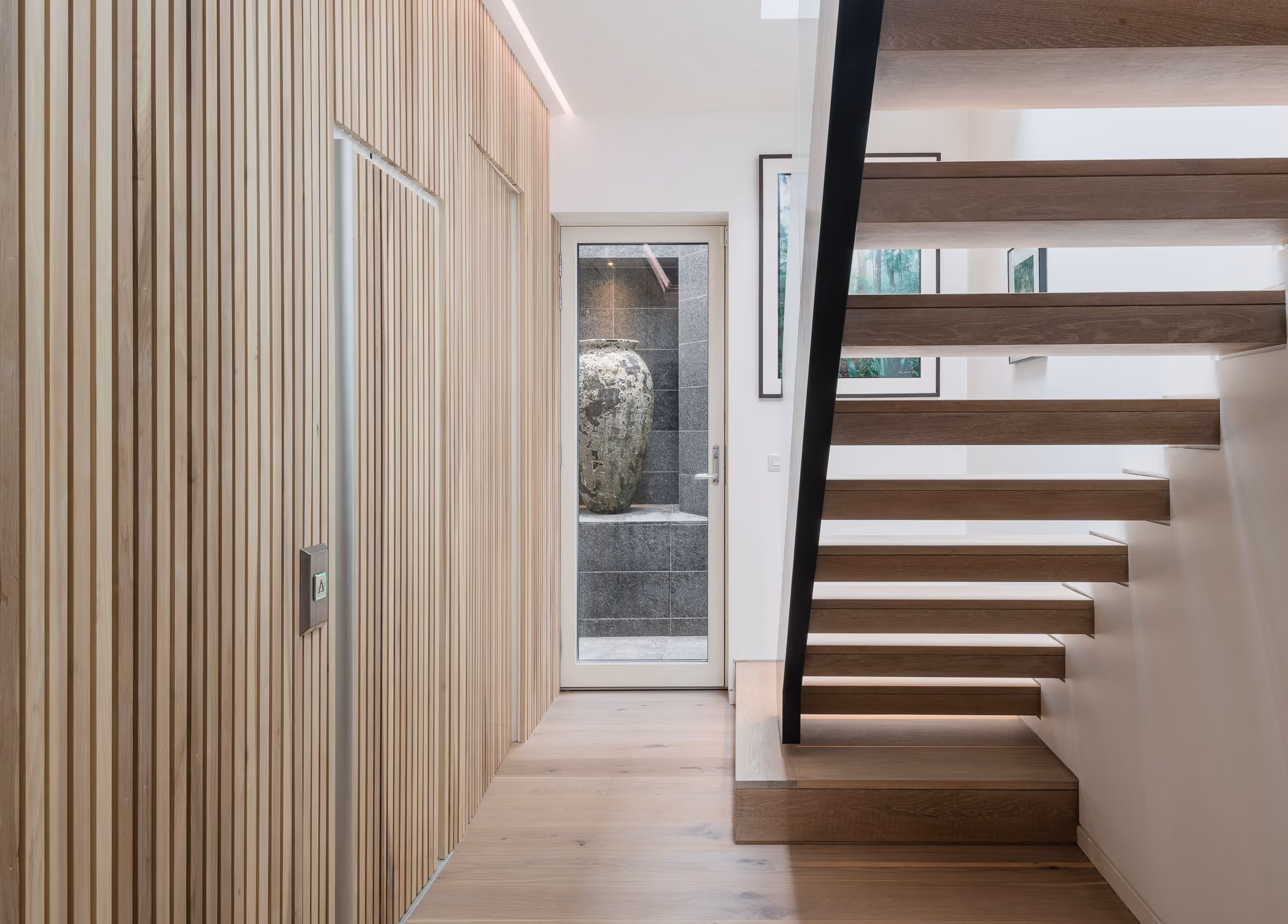Modern interior with floating wooden stairs, vertical wood panel wall, and a glass door with vase outside.