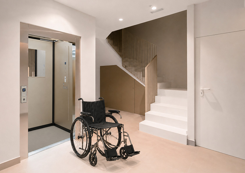 Empty black wheelchair facing an open public elevator inside a modern lobby with beige tiled floor and white stairs with a golden railing.