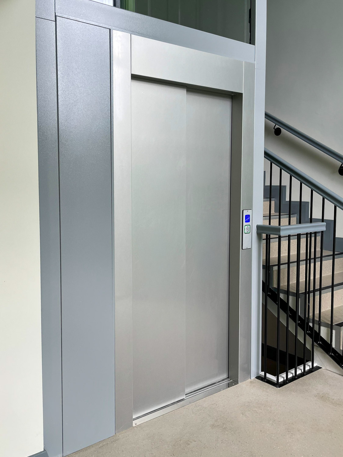 Closed modern stainless steel public elevator with a lit call button panel showing the elevator is on the current floor, located next to a stairway with metal railings and beige steps.