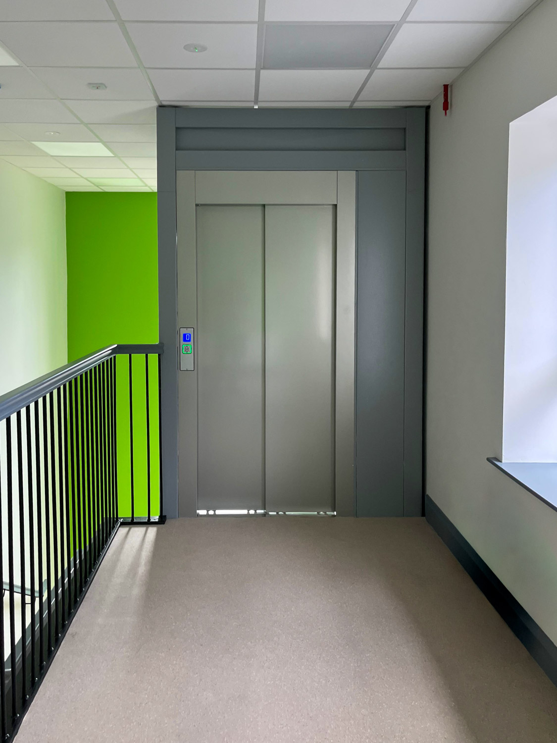 Closed silver elevator doors framed by a gray wall panel with an illuminated control panel showing floor 0, located in a hallway with a black metal railing and a bright green wall on the left, and a window with a white wall on the right.