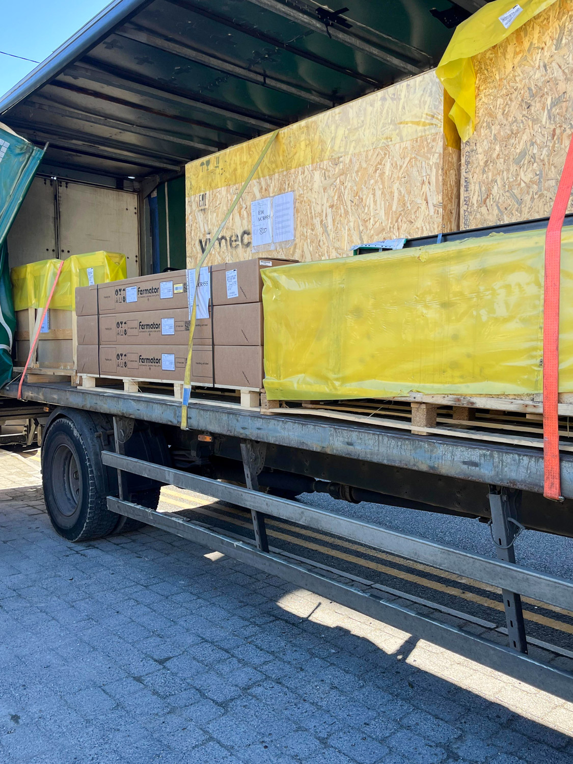 Open flatbed truck loaded with large wooden crates and multiple cardboard boxes labeled Farmator, secured with yellow and orange straps.