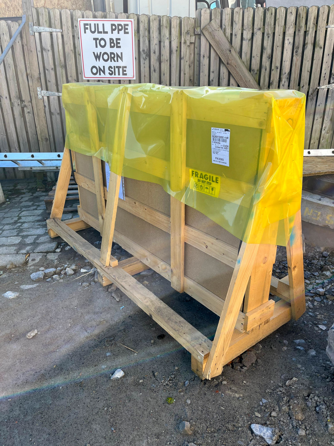 Wooden crate covered with yellow plastic wrap on rough ground near a weathered wooden fence with a sign stating 'FULL PPE TO BE WORN ON SITE', a fragile label is visible on the crate.