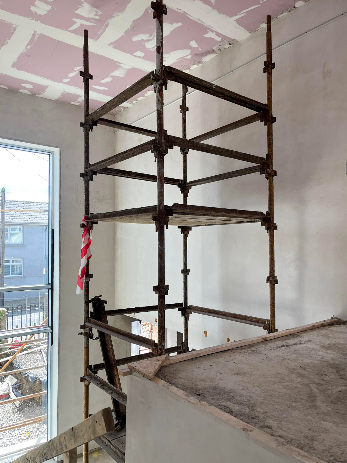 Rusty metal scaffolding structure inside unfinished room with partially plastered white walls and exposed pink ceiling panels, next to a large open window showing an outdoor construction area.