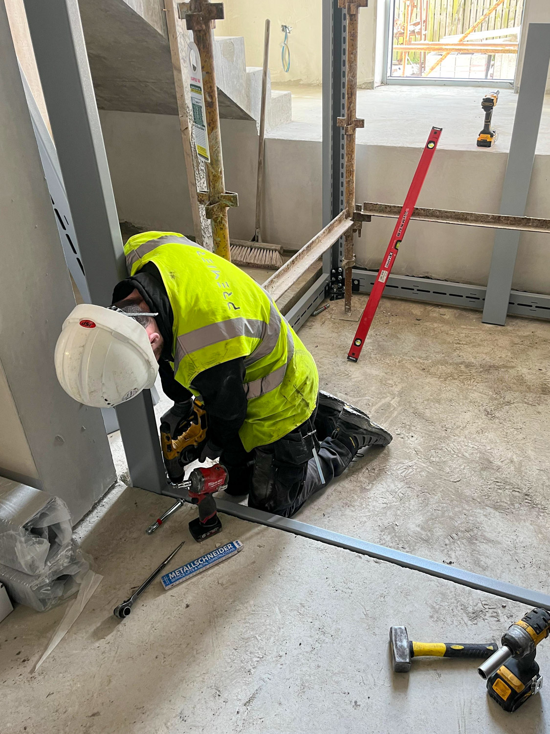 Premier Lifts Ireland engineer wearing a white hard hat and high-visibility yellow vest kneeling on a concrete floor while using a power drill to fasten a metal frame inside a building under construction with tools scattered around.