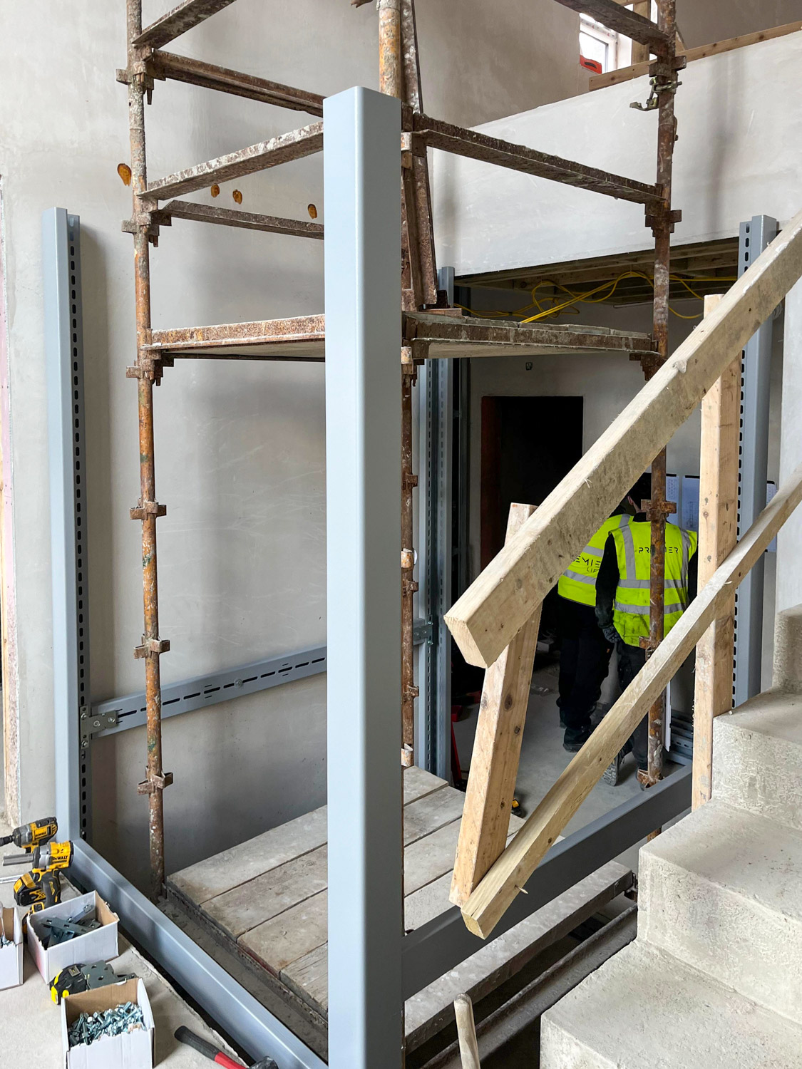 Interior of a building under construction showing a sturdy metal frame installation with scaffolding inside, wooden stairs with a handrail in the foreground, and two Premier Lifts Ireland engineers wearing high-visibility jackets standing inside a dark doorway in the background.