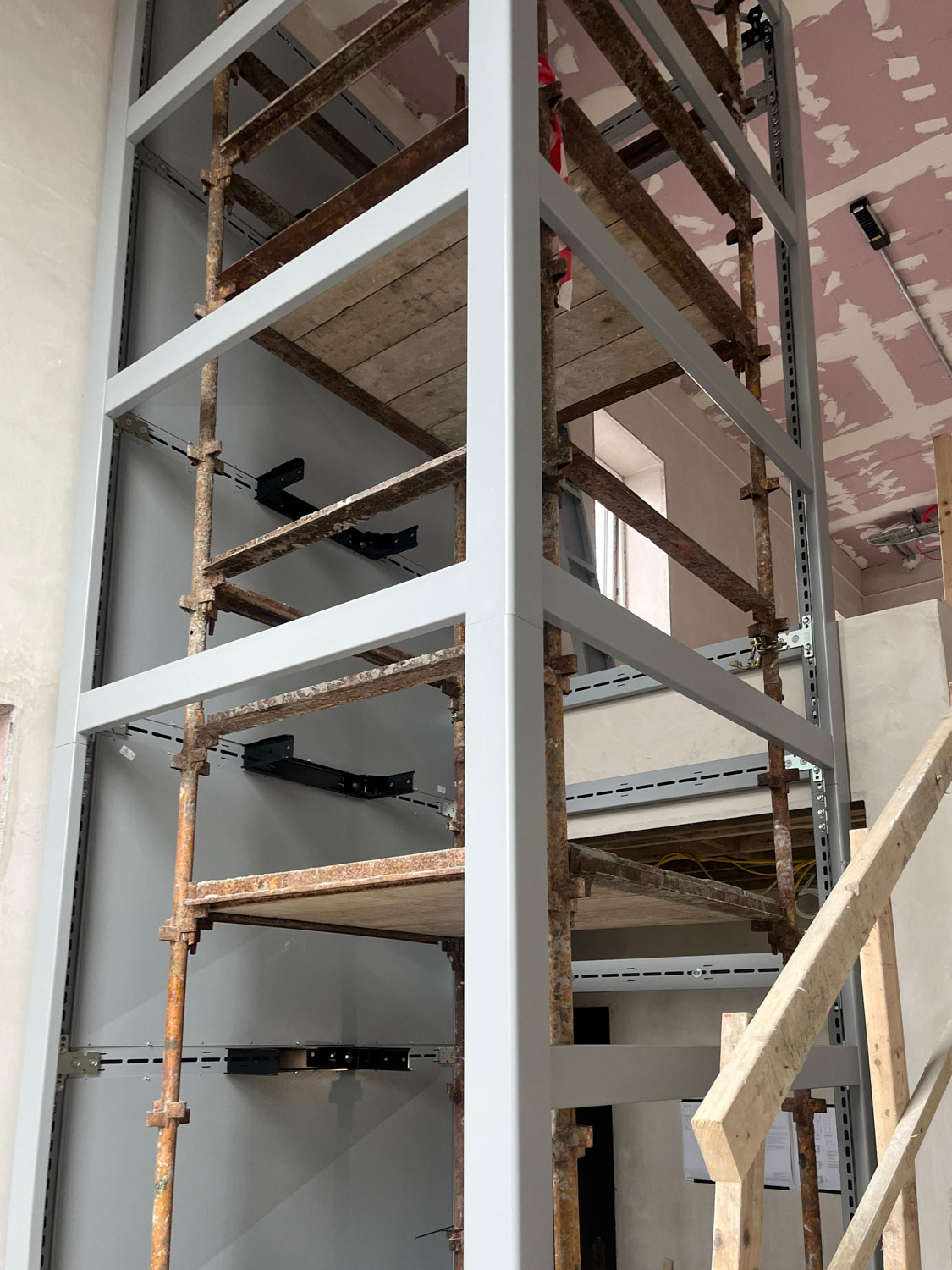 Interior construction scene showing scaffolding built inside a metal frame structure with wooden planks, unfinished walls, and a partially painted ceiling.