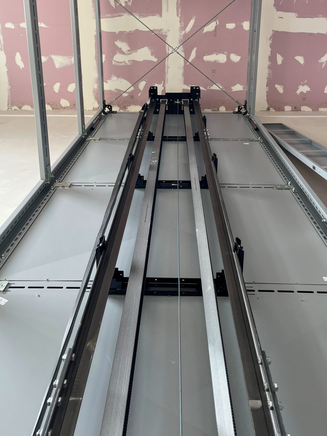 View looking up inside an unfinished elevator shaft showing metal guide rails and cables against walls with exposed drywall.