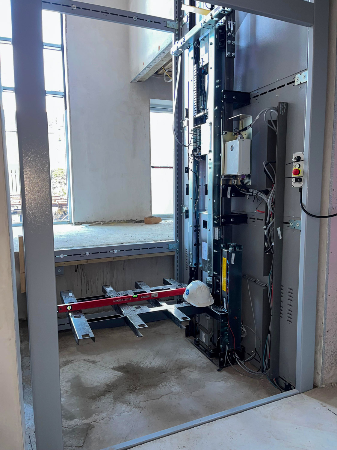 Partially constructed elevator shaft with visible metal framework and electrical components, a red spirit level resting on the metal support, and a white hard hat placed on the framework, inside a building under construction.