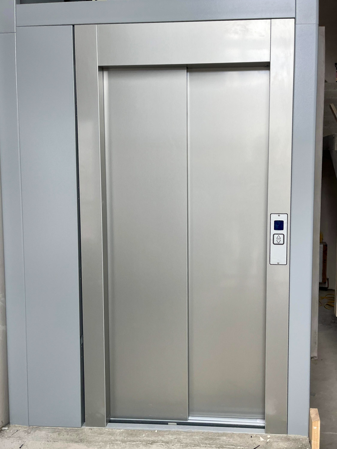 Closed modern silver metal elevator doors set within a gray frame, with an elevator call button panel on the right side.