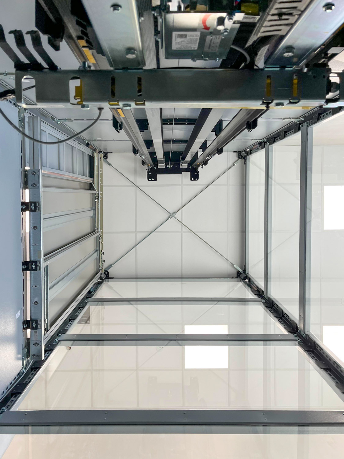 Interior view looking up inside an empty glass elevator shaft showing metal framework, cables, and the ceiling with tiled panels and lights.