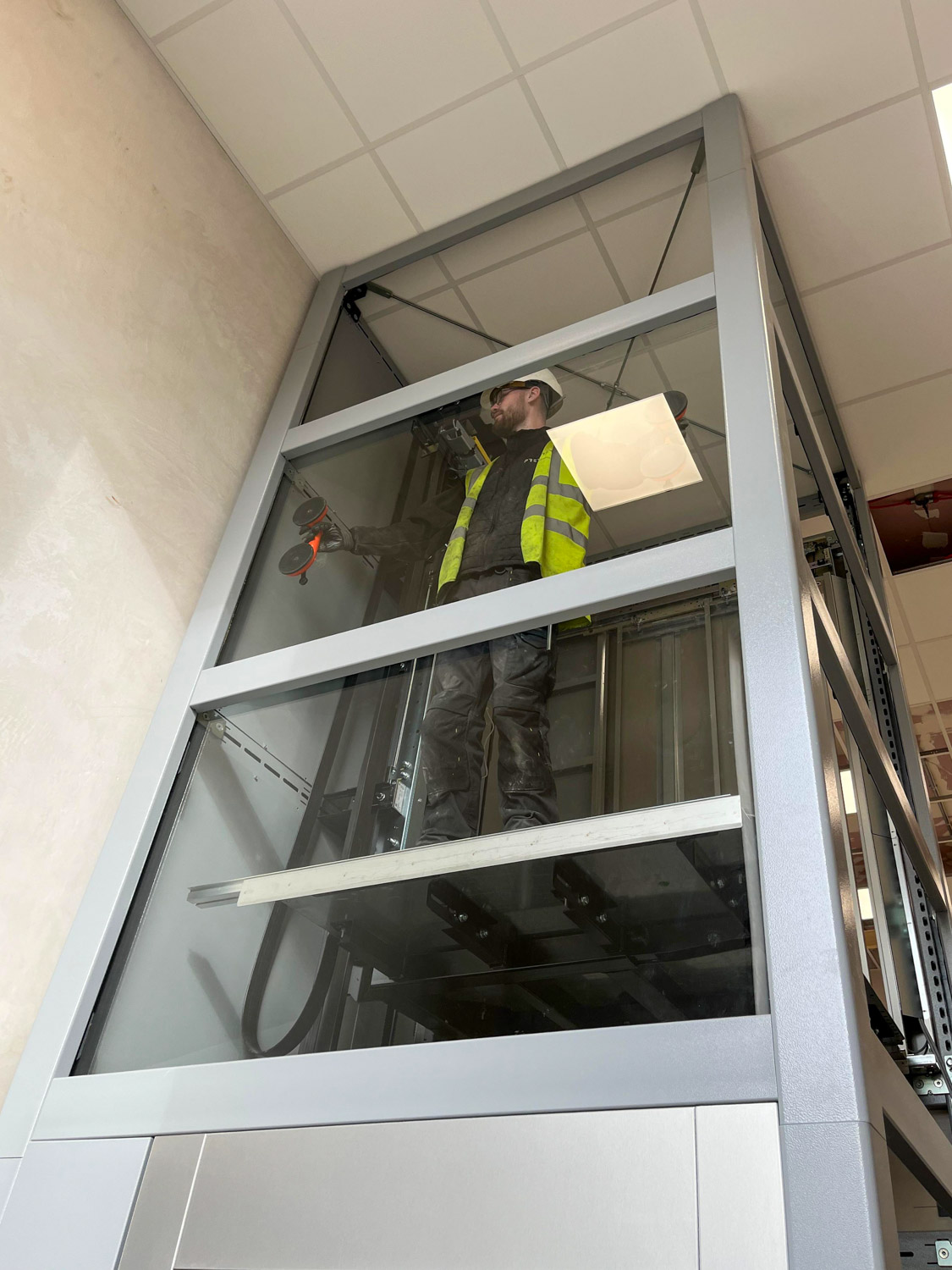 Premier Lifts Ireland engineer wearing a white hard hat, black gloves, and a yellow safety vest inside a glass elevator shaft, holding suction tools and standing on the elevator platform during installation or maintenance.